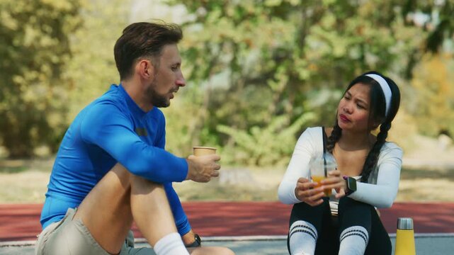 A fit couple takes a refreshing break after their outdoor workout. They sit together, hydrating and conversing in a vibrant setting.