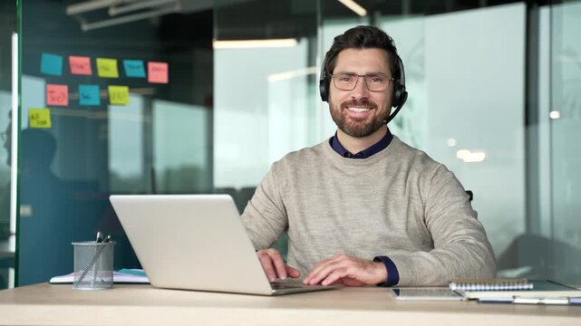 Smiling businessman portrait wearing headset while working on laptop at desk in modern office, then looking at camera and posing. Friendly mature professional working as call center operator, positive