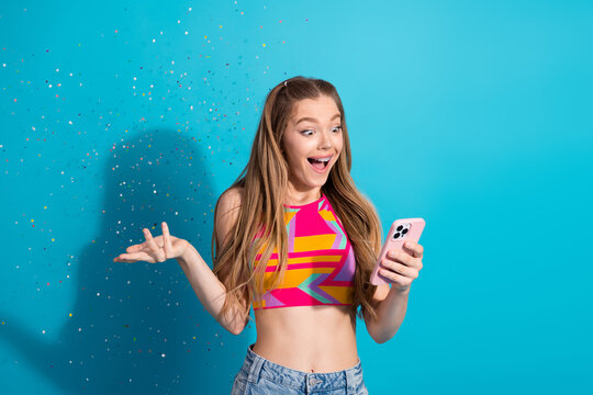 Excited young girl holding smartphone with confetti flying against vibrant blue background, expressing joyous surprise