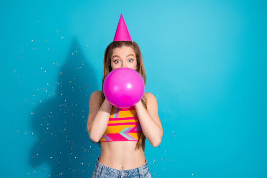 Young girl in colorful top blowing up a pink balloon in celebration, standing against a vibrant blue confetti-filled background
