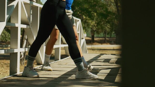 A fit couple performs a stretching routine on a wooden bridge in a sunlit park. They are warming up together before their workout, focusing on flexibility and health.