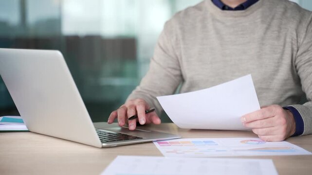 Close up of businessman hand typing on laptop while holding document at desk in modern office. Mature professional comparing data, checking information and reviewing paperwork during focused work.