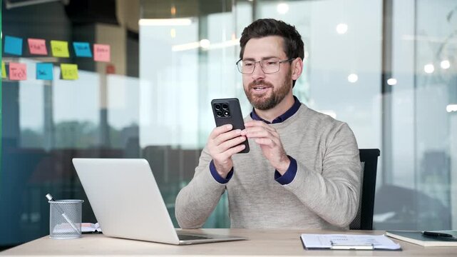 Smiling businessman reacts with pleasant surprise after reading good news on smartphone. Happy mature professional sitting at desk in modern office, satisfied with positive message on mobile phone.