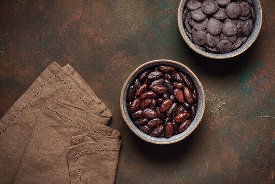Cocoa beans and chocolate buttons in bowls on a dark rustic background, top view, copy space.