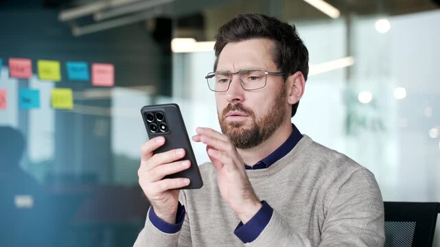 Frustrated businessman facing technical problem on smartphone, close-up. Angry mature professional reacts with irritation while trying to solve mobile connection issue during work in modern office.