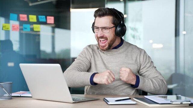 Excited businessman wearing wireless headphones watching sports match on laptop while sitting at desk in modern office. Happy mature professional cheering, celebrating victory with emotional reaction.