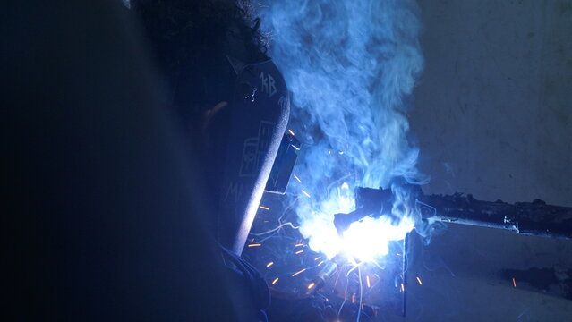 A close-up view of a welding electrode producing a brilliant blue arc and dense smoke while joining metal in a dim industrial environment.