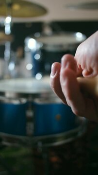 Extreme close up of a musician hand holding a wooden drumstick and tapping a beat, with a drum kit out of focus in the background during a rehearsal or performance in a studio. Vertical video