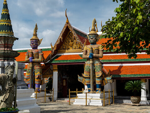 View of The Temple of the Emerald Buddha featuring ornate giant demon guardian statues before a traditional Thai temple with colorful tiled roofs, Krung Thep Maha Nakho, Thailand.