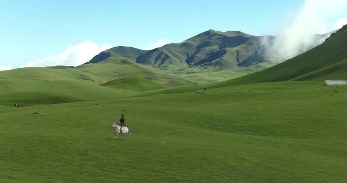 Badimalika region, Bajura, Nepal, aerial shot of a person riding a white horse and running across vast green rolling pastures in Triveni Patan grassland on a bright sunny day