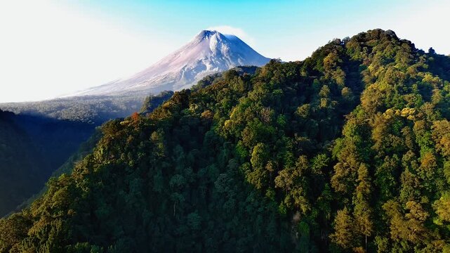 Aerial view of Mount Merapi volcano rising above dense tropical forest, with lush green slopes and clear sky in Pakem, Special Region of Yogyakarta, Indonesia.