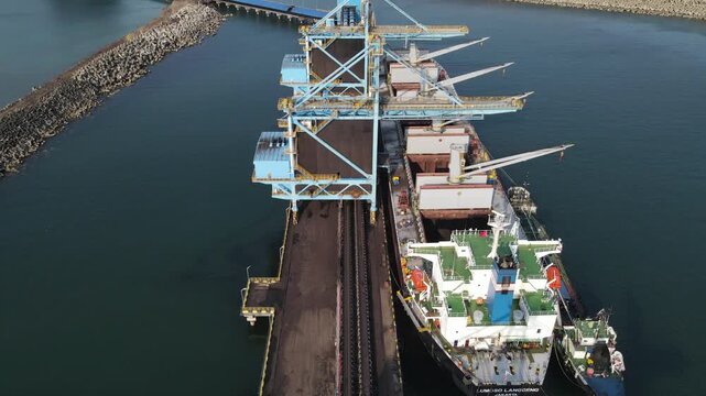 Aerial view of a large cargo ship docked at the port being loaded with coal by an industrial conveyor system in Cilacap, Central Java, Indonesia.