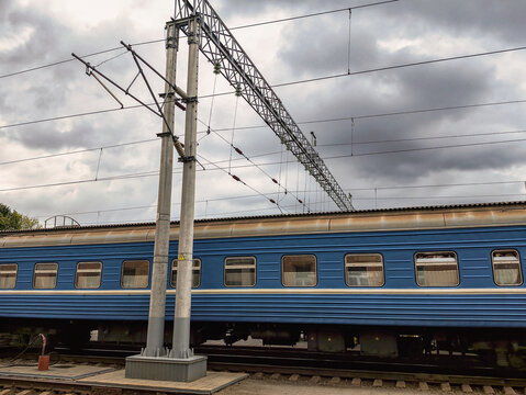 A blue train is parked at a train platform as dark clouds fill the sky. Overhead power lines are visible along the tracks at the station
