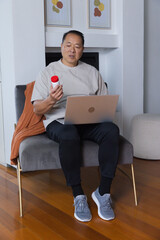Fototapeta premium Senior Asian man sitting in living room, reading white bottle label while using laptop