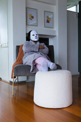 Fototapeta premium Senior African American woman sitting on lounge chair in living room wearing robe and white mask