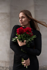 Fototapeta premium Vibrant floral portrait features woman in elegant dark attire holding bunch of red peonies against neutral wall outdoor setting