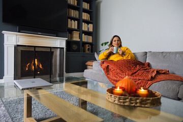 Fototapeta premium Mature African American woman lounging sofa in mustard-top holding blue mug by fireplace