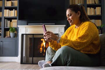 Fototapeta premium African American woman using pink phone at home in mustard top near lit fireplace, copy space