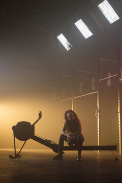 African American woman sitting on rowing erg rail in warehouse gym under skylights