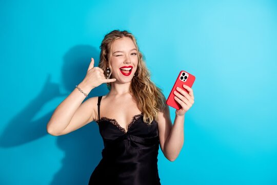 Young woman in black dress holding red smartphone, winking and making a phone hand gesture while smiling against a bright blue background