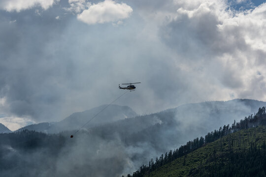 View of a helicopter firefighting in the Fraser Canyon with smoke rising over the forested mountains in Fraser Valley, British Columbia, Canada.