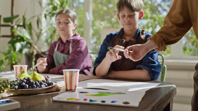 Caucasian teacher passing paintbrush to schoolboy during art masterclass with classmate. Visual works for advertising children education, art school courses and creative development workshops