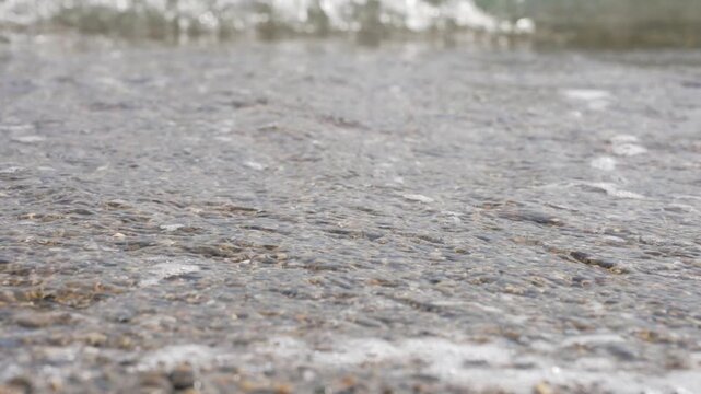 Close up of foamy sea wave washing over pebble beach shore on sunny day, 4k 120fps slow motion static footage