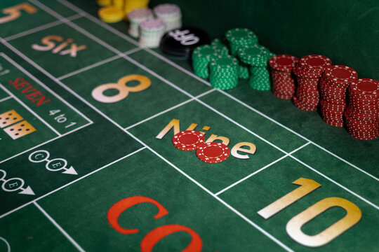 Casino craps table layout with stacks of gambling chips on green felt. Betting grid displays marked numbers and wager positions in close diagonal view.
