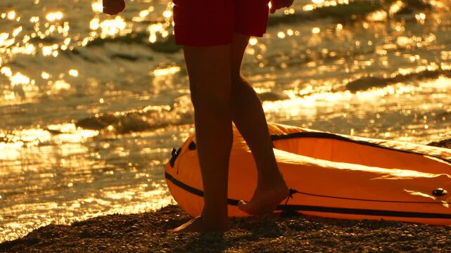 Inflatable yellow raft awaits by golden tide as legs of a person standing on rocky shoreline remain stationary. Low angle side view shot.