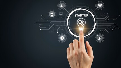 Obraz premium Hand touching lightbulb with startup concept surrounded by digital icons against dark background