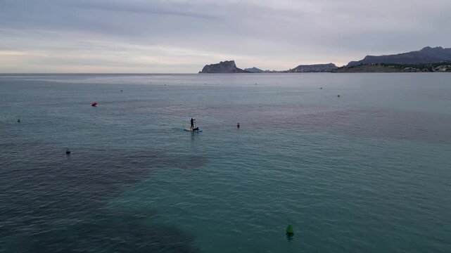 Aerial view of a person paddleboarding on the calm Mediterranean sea with the Penon de Ifach in the distance in Moraira, Valencian Community, Spain.