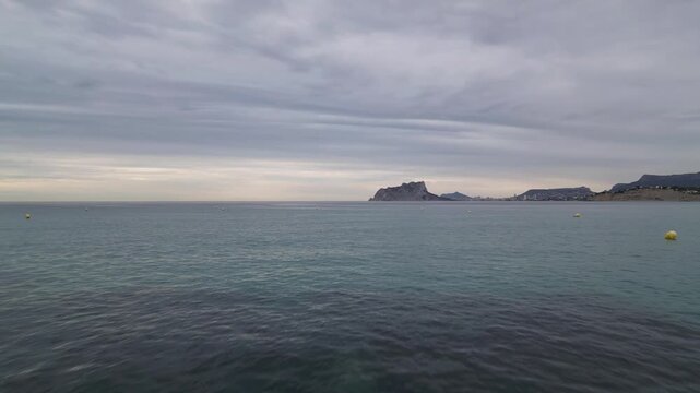 Aerial view of the calm Mediterranean Sea with floating buoys and the distant Penon de Ifach rock formation under a cloudy sky in Moraira, Valencian Community, Spain.