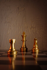 Three golden chess pieces king rook and knight stand on board before textured wall in dramatic warm light with reflections and large negative space above for text © abramov_jora