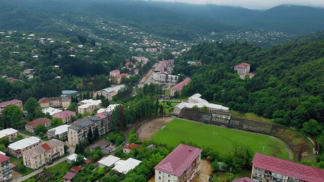 Panoramic view from above the historic resort town of Tkibuli in western Georgia, showcasing Soviet-era architecture, a sports stadium and lush green forests around the urban area