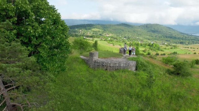Happy young friends waving and rejoicing on top of a hill as the drone flies away, revealing a beautiful green valley with mountains in Georgia's Racha, a lake and a village road on a sunny day with c