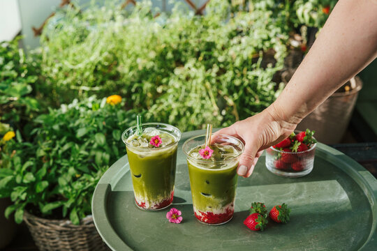 Woman reaching for a refreshing layered strawberry matcha latte on an outdoor table surrounded by lush green plants and fresh strawberries