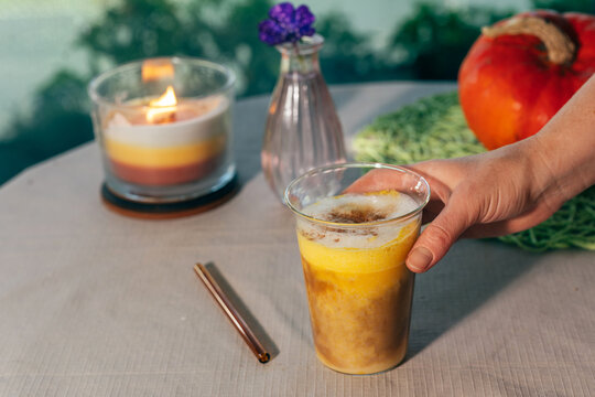 Woman holding a glass of homemade pumpkin spice latte on a cozy outdoor autumn table decorated with a pumpkin, candle and flower