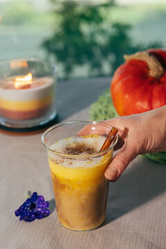 Close-up of a woman holding a creamy pumpkin spice latte on an outdoor table with a pumpkin and glowing candle in the background