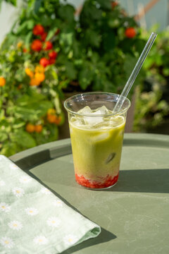 Refreshing layered strawberry matcha latte with ice served on a sunny outdoor table surrounded by colorful flowers and lush greenery