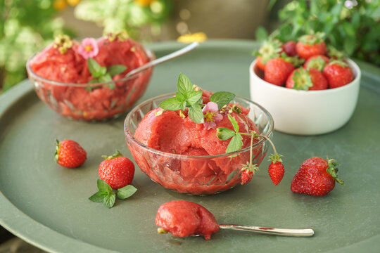 Homemade strawberry sorbet with fresh strawberries, mint and spoons arranged on a sunny outdoor table in a lush summer garden