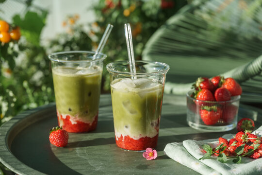 Two colorful layered strawberry matcha lattes served with a bowl of fresh strawberries on a sunny outdoor table surrounded by lush summer greenery