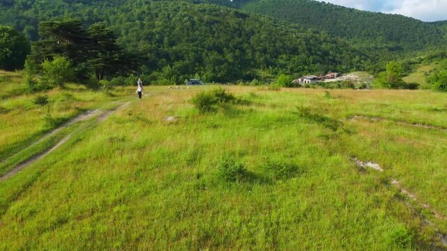 Aerial drone shot captured by two women walking along a dirt road through a bright green meadow while a car with a roof rack stands by, against a backdrop of lush forested mountains racha georgia