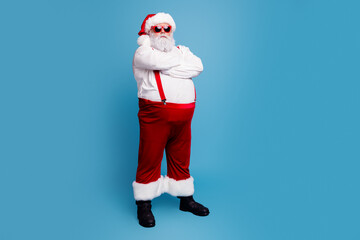 Fototapeta premium Cool Santa Claus standing with arms crossed in red suit and sunglasses on blue background, confident festive Christmas portrait