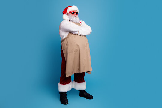 Santa Claus wearing an apron and red sunglasses standing with folded arms on a blue studio background in a playful modern holiday portrait
