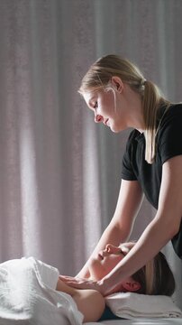 Young girl receives facial and neck massage from therapist. Therapist uses gentle pressure with hands arms visible against soft lighting backdrop woman