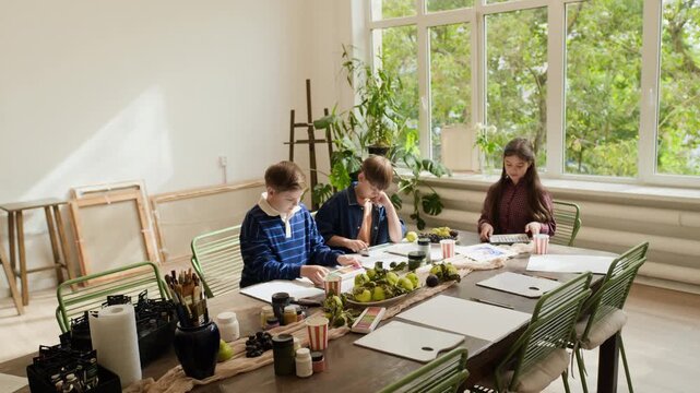 Caucasian children attending art master class, girl showing colorful painting to boys at creative workshop. Footage suitable for youth education campaigns and art school promotion