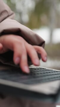 Close up shows woman hands actively pressing and gliding over black keycaps of silver laptop. soft focus background suggests outdoor scene with muted greenery while natural light illuminates