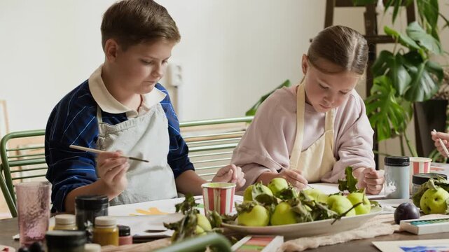 Caucasian boy and girl sitting at table, painting still life during art master class. Footage suitable for creative education campaigns, children workshop advertising, and parenting blogs