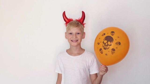 Kids halloween costume for party store ads, smiling boy in red devil horns and face paint holds orange skull balloon on light background. Costume party and trick or treat fun, boy looks at camera