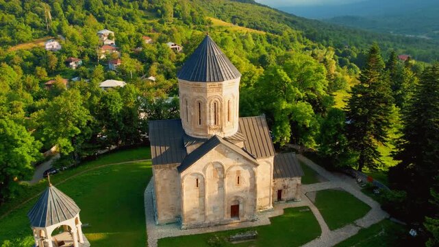 Aerial drone footage circling the historic nikortsminda cathedral, a unesco tentative site in georgia, showing its detailed stone architecture against a beautiful sunlit mountain landscape